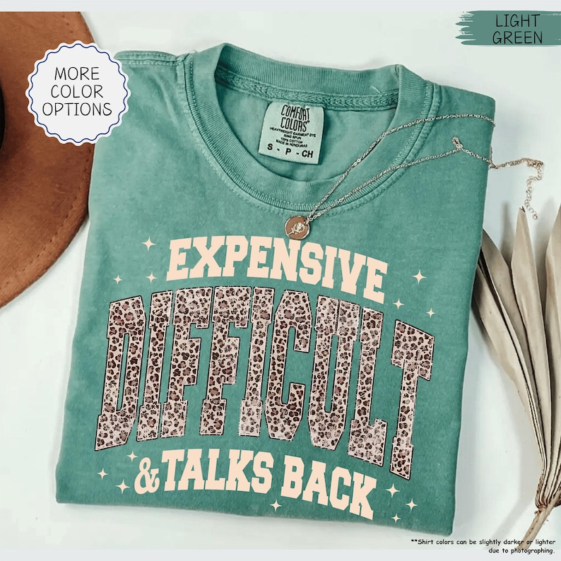 May include: A light green Comfort Colors t-shirt with the words "EXPENSIVE DIFFICULT & TALKS BACK" in a combination of pink and leopard print. A silver necklace with a pendant is draped across the shirt. The shirt is folded.