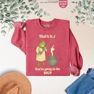 Pu&ograve; includere: Felpa rosso scarlatto melange con un'illustrazione di uno chef rana. La rana sta mescolando una pentola di zuppa con il testo "That's it. You're going in the SOUP" stampato sotto.