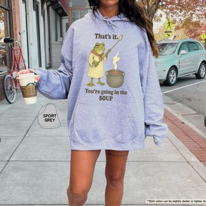 Pu&ograve; includere: Felpa con cappuccio grigio sportivo con un'illustrazione di rana e la scritta "That's it. You're going in the SOUP." La rana tiene un bastone sopra una pentola. La persona indossa una tazza di caff&egrave;.