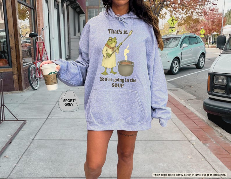 Pu&ograve; includere: Felpa con cappuccio grigio sportivo con un'illustrazione di rana e la scritta "That's it. You're going in the SOUP." La rana tiene un bastone sopra una pentola. La persona indossa una tazza di caff&egrave;.