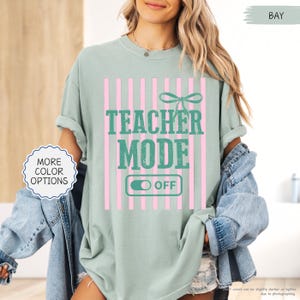 May include: A light green t-shirt with the words "TEACHER MODE OFF" in teal, set against a pink striped background. A bow graphic is above the text. The shirt also has a "MORE COLOR OPTIONS" button.