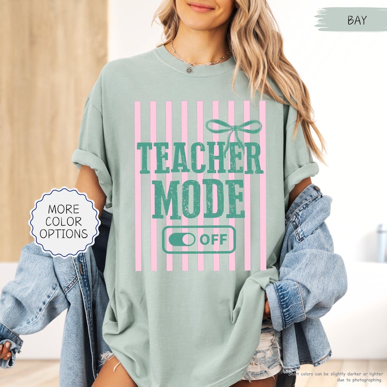 May include: A light green t-shirt with the words "TEACHER MODE OFF" in teal, set against a pink striped background. A bow graphic is above the text. The shirt also has a "MORE COLOR OPTIONS" button.