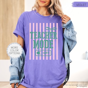 May include: A purple t-shirt with the text "TEACHER MODE OFF" in green, set against a pink and white striped background. The shirt has short sleeves and a relaxed fit. The image also includes a denim jacket.
