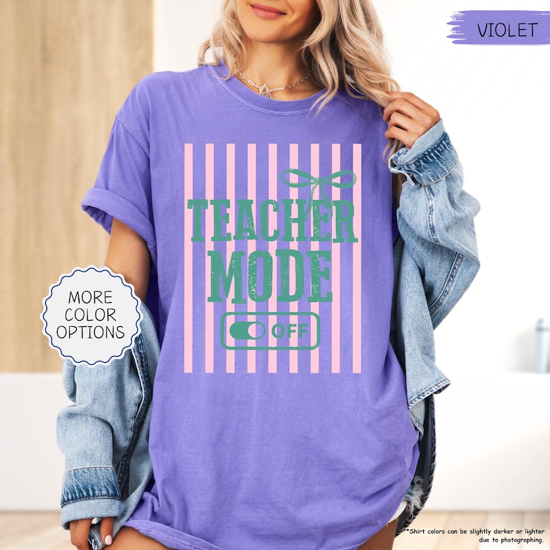 May include: A purple t-shirt with the text "TEACHER MODE OFF" in green, set against a pink and white striped background. The shirt has short sleeves and a relaxed fit. The image also includes a denim jacket.