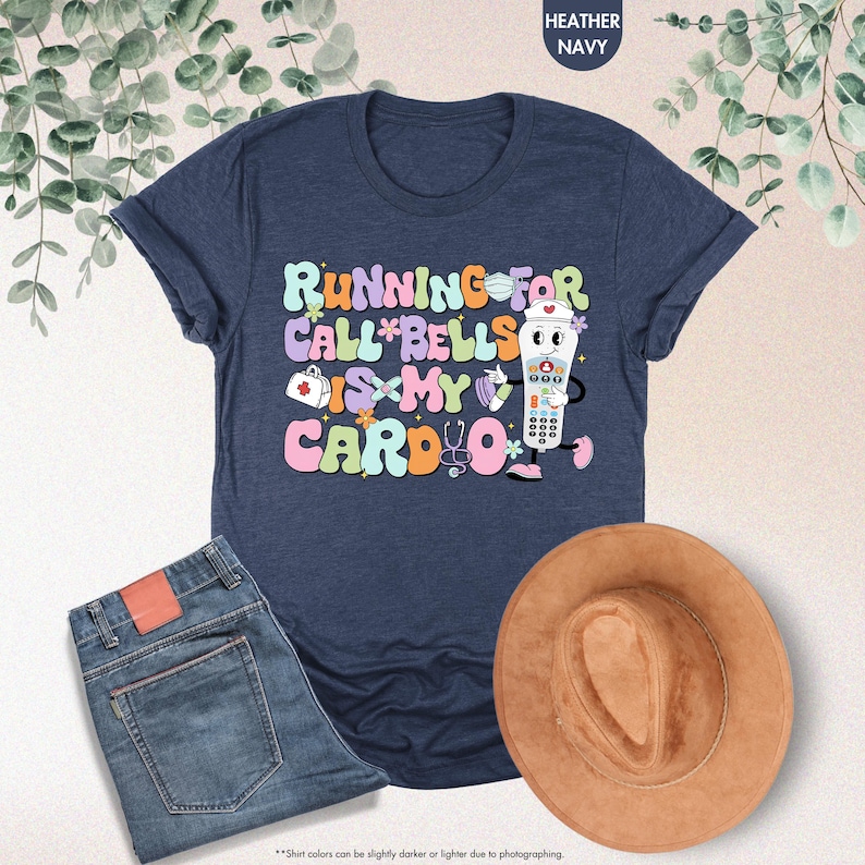 May include: A navy blue t-shirt with a graphic of a cartoon nurse holding a medical alert button. The text on the shirt reads "Running for Call Bells is my Cardio".