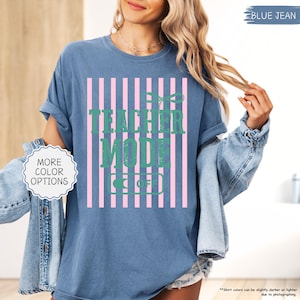 May include: Blue jean-colored t-shirt with the text "TEACHER MODE OFF" in green, set against a pink and white striped background. The shirt has rolled-up sleeves and a relaxed fit. The words "MORE COLOR OPTIONS" are in a white circle.