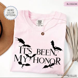 Pu&ograve; includere: T-shirt rosa chiaro con la scritta "IT'S BEEN. MY HONOR" e grafica di pipistrelli neri. La maglietta ha scollo tondo e maniche corte. Una delicata collana con un ciondolo &egrave; appoggiata sulla maglietta.
