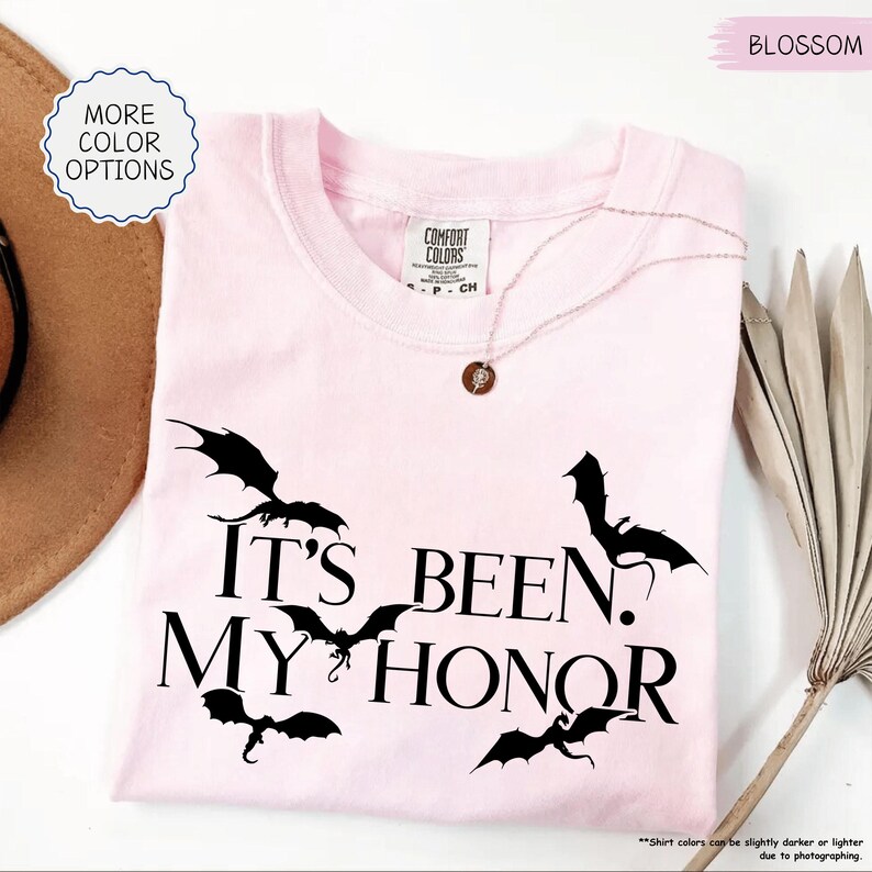 Pu&ograve; includere: T-shirt rosa chiaro con la scritta "IT'S BEEN. MY HONOR" e grafica di pipistrelli neri. La maglietta ha scollo tondo e maniche corte. Una delicata collana con un ciondolo &egrave; appoggiata sulla maglietta.