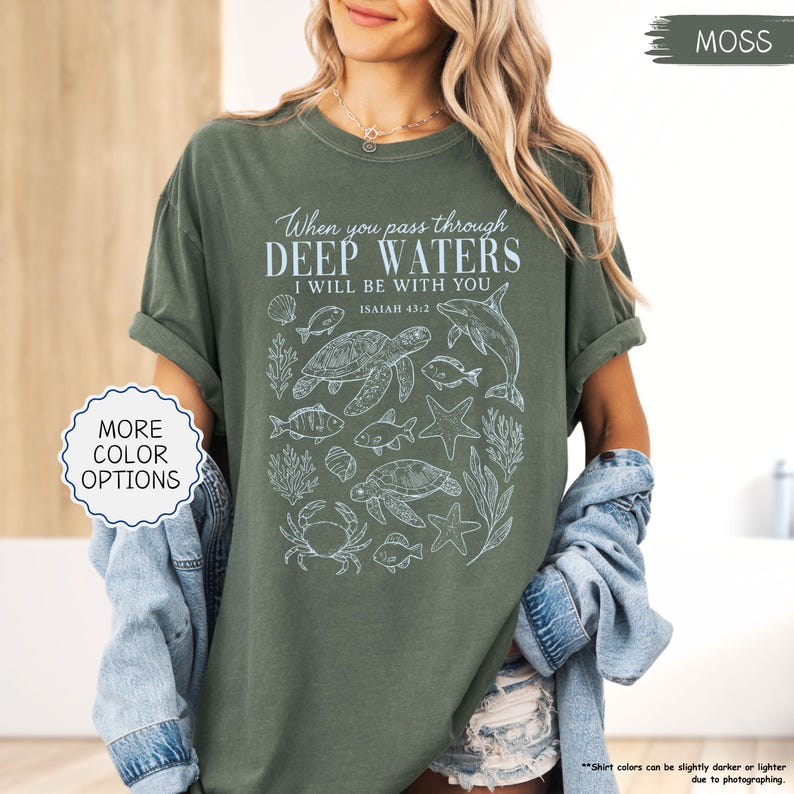 May include: Moss green t-shirt with the text "When you pass through deep waters, I will be with you" and Isaiah 43:2. The shirt features a white graphic of sea creatures. The shirt is a relaxed fit.