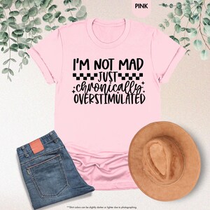 Op de afbeelding: Lichtroze t-shirt met de zwarte tekst "I'M NOT MAD JUST chronically OVERSTIMULATED". De afbeelding toont ook een blauwe jeans en een bruine hoed. Het t-shirt is lichtroze van kleur.