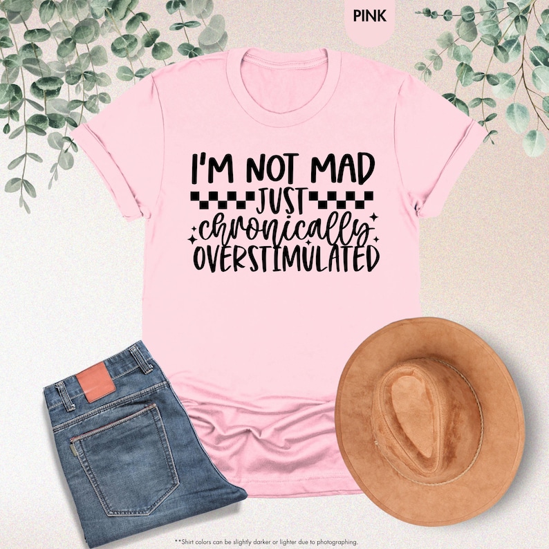 Op de afbeelding: Lichtroze t-shirt met de zwarte tekst "I'M NOT MAD JUST chronically OVERSTIMULATED". De afbeelding toont ook een blauwe jeans en een bruine hoed. Het t-shirt is lichtroze van kleur.