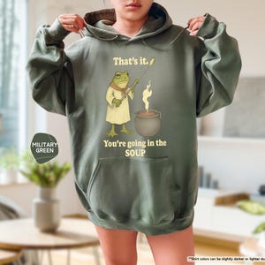 Pu&ograve; includere: Felpa con cappuccio verde militare con una grafica di una rana che tiene un mestolo sopra una pentola di zuppa. Il testo sulla felpa recita: "That's it. You're going in the SOUP."