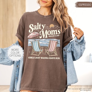 May include: Espresso-colored t-shirt with a beach-themed graphic. The design features the text "Salty Moms - Beach Club" above two beach chairs, an umbrella, and the phrase "Girls Just Wanna Have Sun."