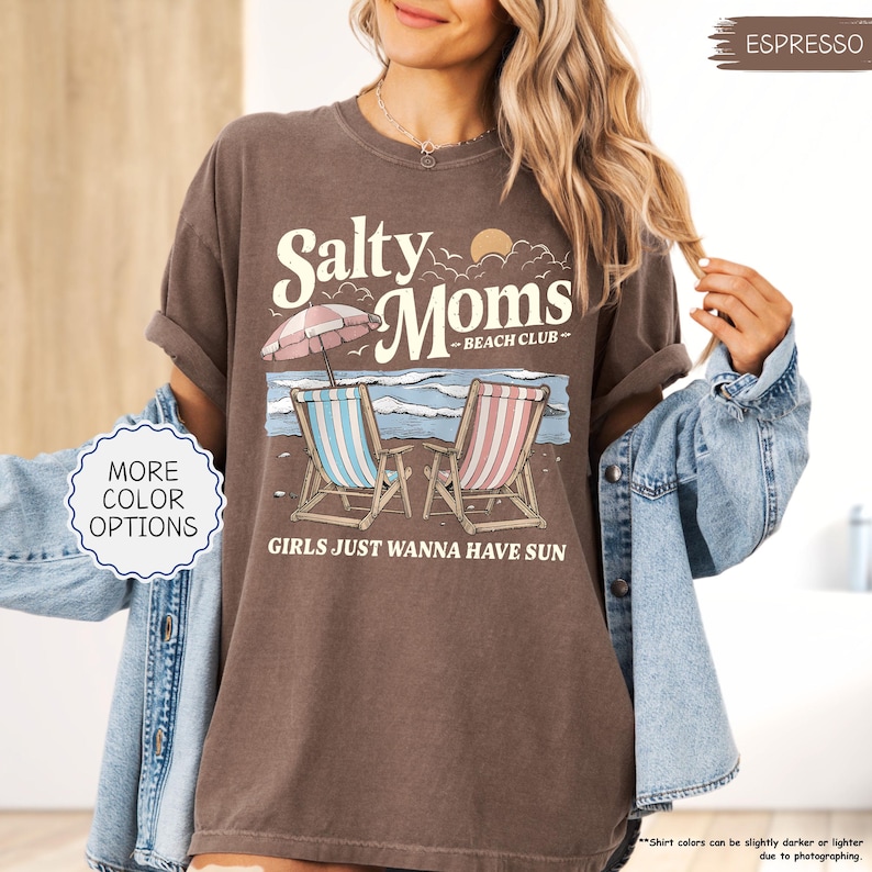 May include: Espresso-colored t-shirt with a beach-themed graphic. The design features the text "Salty Moms - Beach Club" above two beach chairs, an umbrella, and the phrase "Girls Just Wanna Have Sun."
