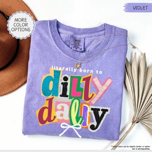 K&ouml;nnte beinhalten: Ein violettes Comfort Colors T-Shirt mit dem Satz "literally born to dilly dally" in farbenfrohem, geschichtetem Text. Eine goldene Halskette und ein brauner Hut sind sichtbar. Das Shirt ist in der Gr&ouml;&szlig;e Small, Petite und Child.