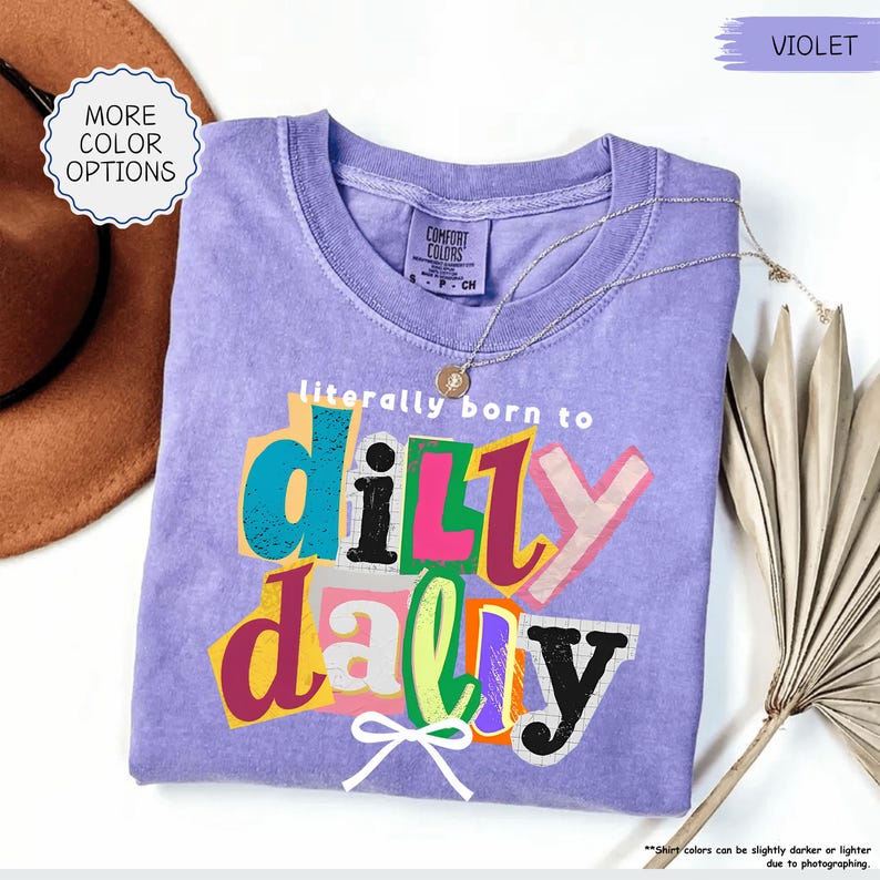 K&ouml;nnte beinhalten: Ein violettes Comfort Colors T-Shirt mit dem Satz "literally born to dilly dally" in farbenfrohem, geschichtetem Text. Eine goldene Halskette und ein brauner Hut sind sichtbar. Das Shirt ist in der Gr&ouml;&szlig;e Small, Petite und Child.