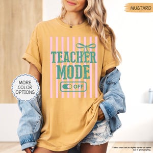 May include: Mustard-colored t-shirt with the text "TEACHER MODE OFF" in teal and pink. The design includes a bow and vertical pink stripes. The shirt has rolled-up sleeves and is worn with a denim jacket.