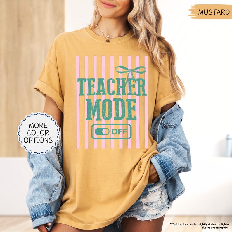 May include: Mustard-colored t-shirt with the text "TEACHER MODE OFF" in teal and pink. The design includes a bow and vertical pink stripes. The shirt has rolled-up sleeves and is worn with a denim jacket.