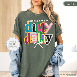 K&ouml;nnte beinhalten: Moosgr&uuml;nes T-Shirt mit dem Text "literally born to dilly dally" in einem farbenfrohen Design im Collage-Stil. Das Shirt hat hochgekrempelte &Auml;rmel und eine lockere Passform. Ein Knopf mit "MORE COLOR OPTIONS" befindet sich links.