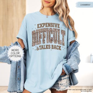 May include: Light blue t-shirt with the words "EXPENSIVE, DIFFICULT, & TALKS BACK" in a brown leopard print font. The shirt has rolled-up sleeves and is paired with a denim jacket and shorts. The words "MORE COLOR OPTIONS" are also visible.