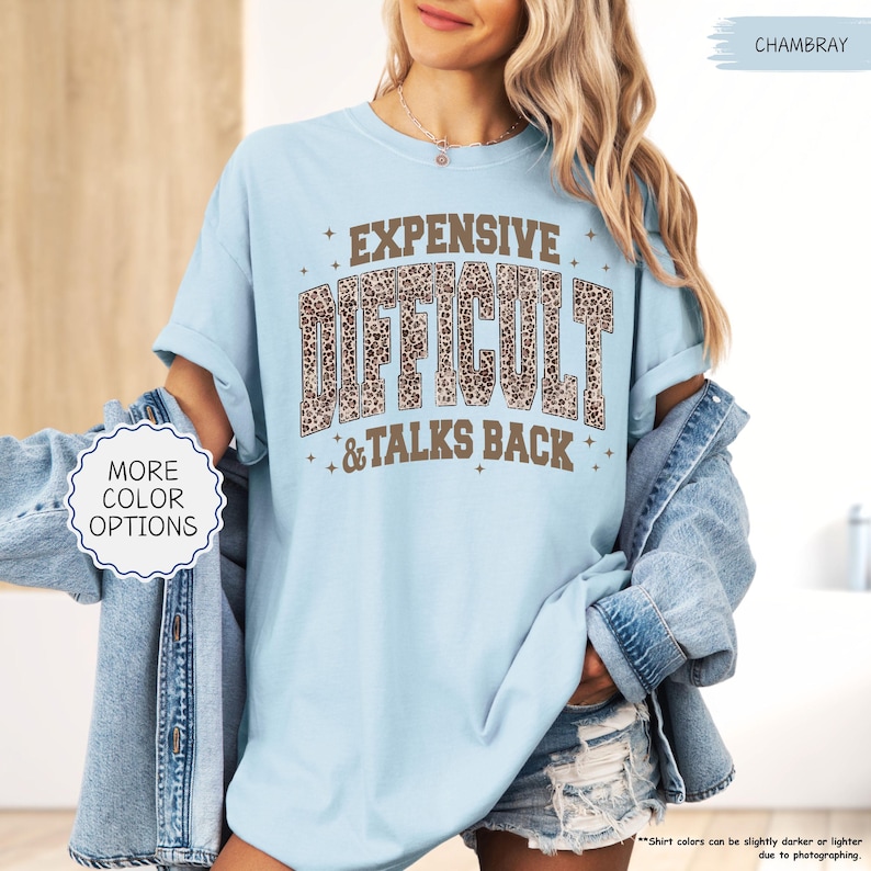 May include: Light blue t-shirt with the words "EXPENSIVE, DIFFICULT, & TALKS BACK" in a brown leopard print font. The shirt has rolled-up sleeves and is paired with a denim jacket and shorts. The words "MORE COLOR OPTIONS" are also visible.