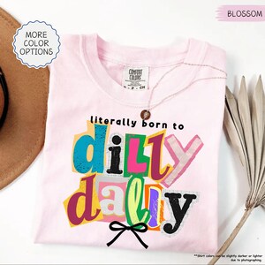 K&ouml;nnte beinhalten: Ein hellrosa T-Shirt mit dem Schriftzug "literally born to dilly dally" in bunten Blockbuchstaben. Das T-Shirt hat einen Rundhalsausschnitt und kurze &Auml;rmel. Eine zarte Halskette liegt auf dem Shirt. Das Shirt ist mit "Blossom" gekennzeichnet.