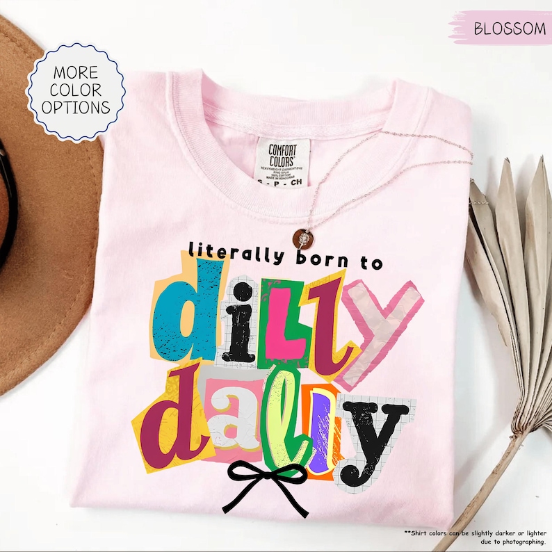 K&ouml;nnte beinhalten: Ein hellrosa T-Shirt mit dem Schriftzug "literally born to dilly dally" in bunten Blockbuchstaben. Das T-Shirt hat einen Rundhalsausschnitt und kurze &Auml;rmel. Eine zarte Halskette liegt auf dem Shirt. Das Shirt ist mit "Blossom" gekennzeichnet.