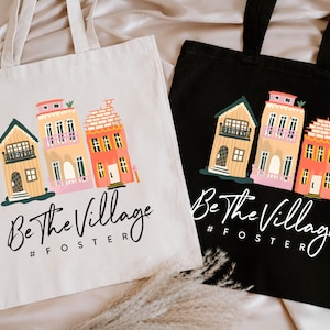 May include: Two tote bags, one cream and one black, each featuring a colorful illustration of three houses. The text "Be The Village #FOSTER" is printed below the houses in a script font.