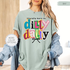K&ouml;nnte beinhalten: Hellgr&uuml;nes T-Shirt mit dem Text "literally born to dilly dally" in bunten, geschichteten Buchstaben. Das Shirt hat hochgekrempelte &Auml;rmel und eine lockere Passform. Eine kleine Schleifengrafik befindet sich unter dem Text. Die Worte "MORE COLOR OPTIONS" stehen in einem Kreis links.