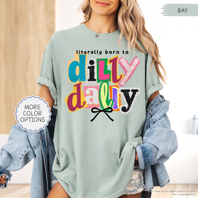 K&ouml;nnte beinhalten: Hellgr&uuml;nes T-Shirt mit dem Text "literally born to dilly dally" in bunten, geschichteten Buchstaben. Das Shirt hat hochgekrempelte &Auml;rmel und eine lockere Passform. Eine kleine Schleifengrafik befindet sich unter dem Text. Die Worte "MORE COLOR OPTIONS" stehen in einem Kreis links.