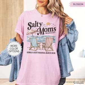 May include: Pink t-shirt with the text "Salty Moms - Beach Club" and the phrase "Girls Just Wanna Have Sun." The graphic features beach chairs, an umbrella, and a sun. The shirt is a light pink color.