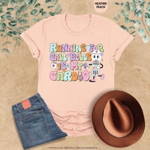 May include: A pink t-shirt with the text "Running for call bells is my cardio" in colorful letters. A cartoon illustration of a call button with a nurse's cap and a stethoscope is next to the text.