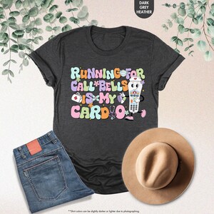 May include: A dark heather grey t-shirt with a graphic of a nurse holding a call button. The text on the shirt reads "Running for call bells is my cardio".