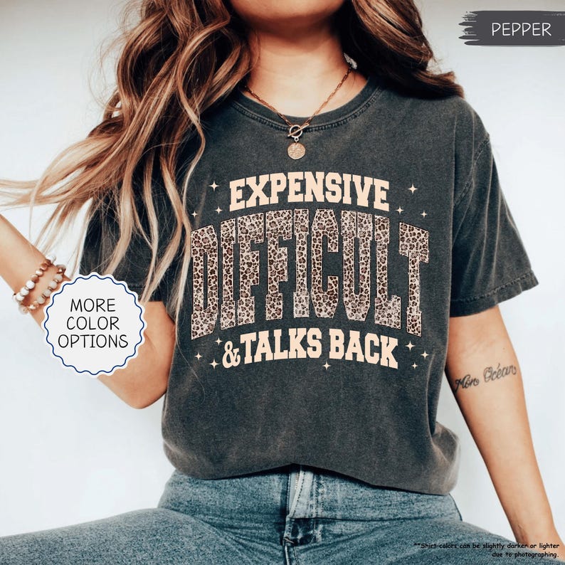 May include: Dark gray t-shirt with the text "EXPENSIVE DIFFICULT & TALKS BACK" in a leopard print design. The shirt has a vintage, worn look. The words "MORE COLOR OPTIONS" are also visible.