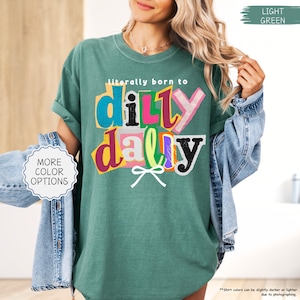 K&ouml;nnte beinhalten: Ein hellgr&uuml;nes T-Shirt mit dem Schriftzug "literally born to dilly dally" in einem farbenfrohen, ausgeschnittenen Stil. Das Shirt hat kurze &Auml;rmel und eine lockere Passform. Die Worte "MORE COLOR OPTIONS" stehen in einem wei&szlig;en Kreis.