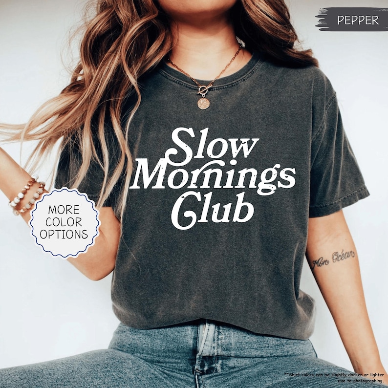 May include: A charcoal grey t-shirt featuring the white text "Slow Mornings Club". The shirt has a faded, vintage look. The image also includes a graphic that says "More Color Options".