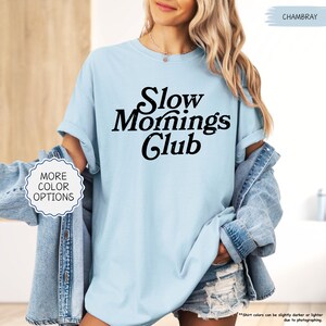 May include: A pale blue t-shirt featuring the words "Slow Mornings Club" in black. The shirt has rolled-up sleeves. The image also shows a denim jacket and shorts. The text "More Color Options" is also visible.