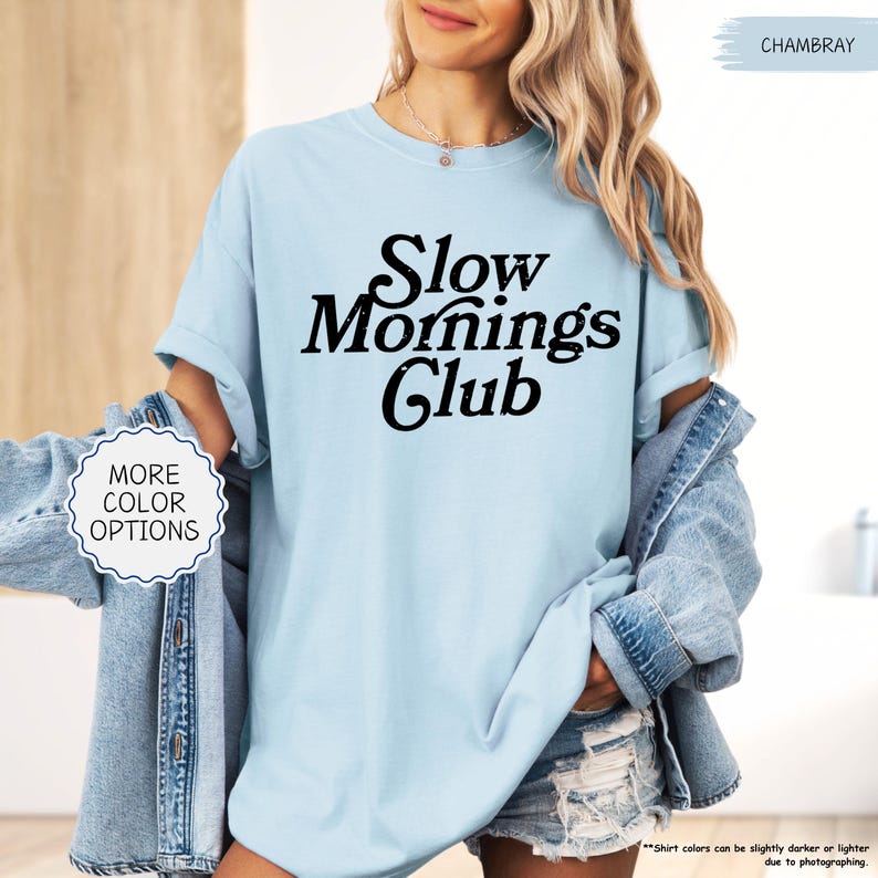 May include: A pale blue t-shirt featuring the words "Slow Mornings Club" in black. The shirt has rolled-up sleeves. The image also shows a denim jacket and shorts. The text "More Color Options" is also visible.