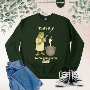 Pu&ograve; includere: Felpa verde foresta con una grafica di un ranocchio chef che tiene un cucchiaio sopra una pentola di zuppa. Il testo recita: "That's it. You're going in the SOUP."
