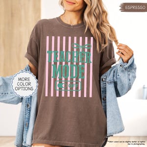 May include: Espresso-colored t-shirt with the text "TEACHER MODE OFF" in teal, set against a pink striped background. The shirt has rolled-up sleeves and is part of a collection with more color options.