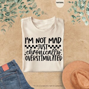 Op de afbeelding: Cr&egrave;mekleurig T-shirt met de zwarte tekst "I'M NOT MAD JUST chronically OVERSTIMULATED". Het shirt ligt plat met een spijkerbroek en een bruine hoed zichtbaar in de afbeelding.