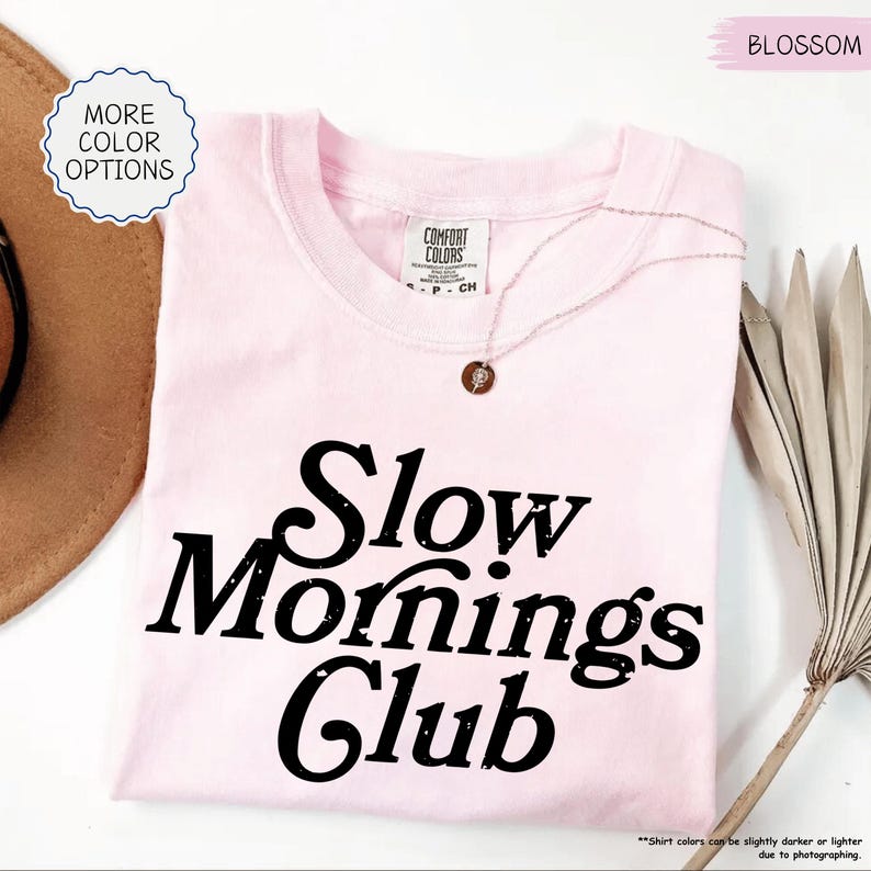 May include: A pale pink t-shirt with the words "Slow Mornings Club" printed in black. The shirt has a crew neck and short sleeves. A delicate necklace with a small pendant rests on the shirt. The words "More Color Options" are in a circle on the top left.