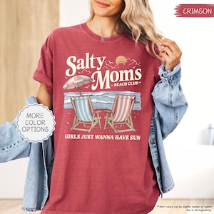 May include: Crimson-colored t-shirt with the text "Salty Moms - Beach Club" and the phrase "Girls Just Wanna Have Sun." The graphic features beach chairs, an umbrella, and ocean waves. The shirt is a relaxed fit.