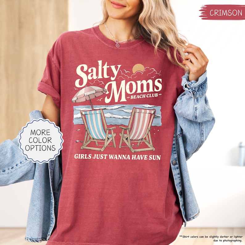 May include: Crimson-colored t-shirt with the text "Salty Moms - Beach Club" and the phrase "Girls Just Wanna Have Sun." The graphic features beach chairs, an umbrella, and ocean waves. The shirt is a relaxed fit.