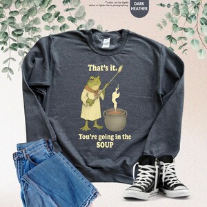 Pu&ograve; includere: Felpa grigio scuro con un'illustrazione di uno chef rana che tiene un cucchiaio sopra una pentola di zuppa. Il testo recita "That's it. You're going in the SOUP." Sono visibili anche jeans e scarpe da ginnastica.