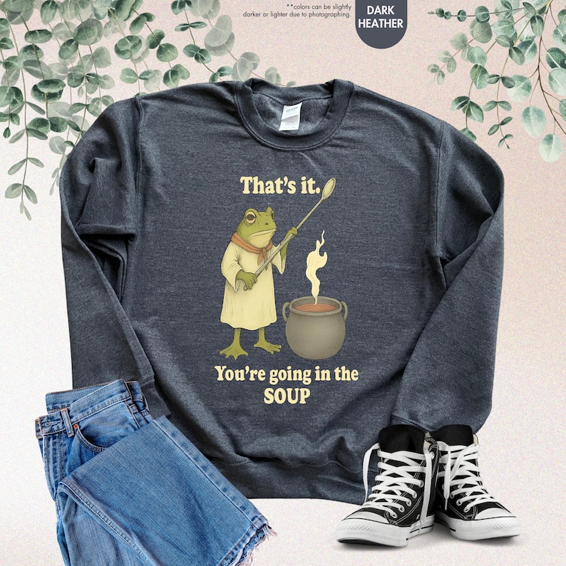 Pu&ograve; includere: Felpa grigio scuro con un'illustrazione di uno chef rana che tiene un cucchiaio sopra una pentola di zuppa. Il testo recita "That's it. You're going in the SOUP." Sono visibili anche jeans e scarpe da ginnastica.