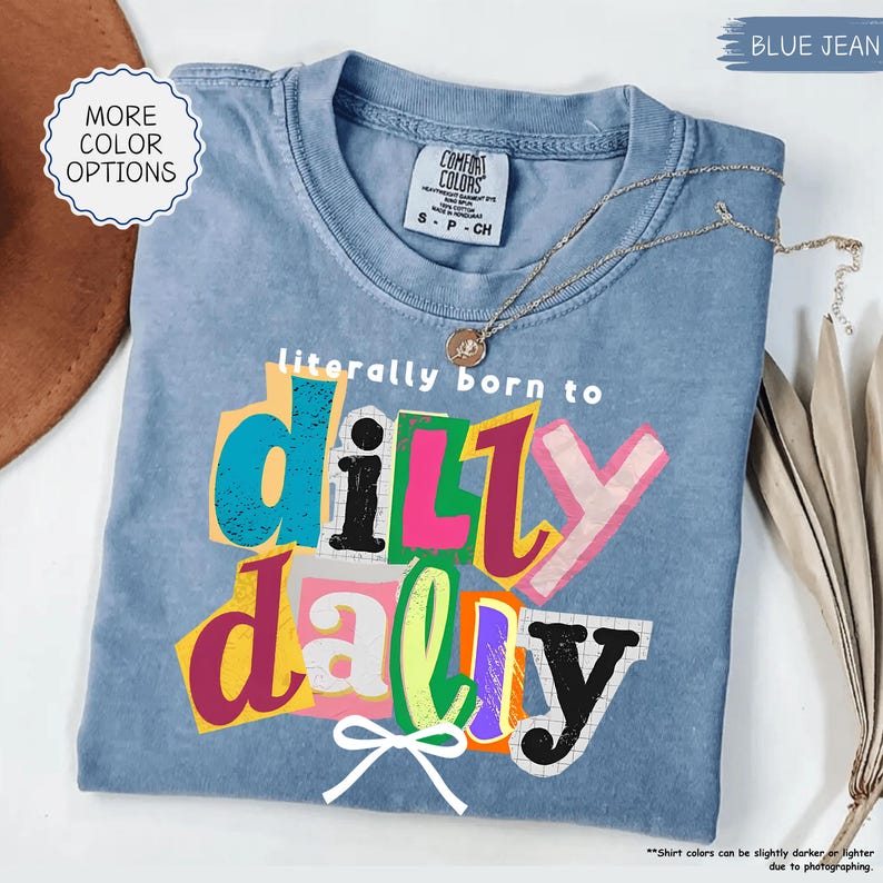K&ouml;nnte beinhalten: Ein blaues Jeans-T-Shirt mit dem Schriftzug "literally born to dilly dally" in bunten, ausgeschnittenen Buchstaben. Eine goldene Halskette und eine wei&szlig;e Schleife sind ebenfalls sichtbar. Das Shirt ist von der Marke Comfort Colors, Gr&ouml;&szlig;e S-P-CH.