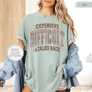 May include: A light green t-shirt with the words "EXPENSIVE DIFFICULT & TALKS BACK" in brown and leopard print lettering. The shirt has rolled-up sleeves and a relaxed fit. The words "MORE COLOR OPTIONS" are in a white circle.
