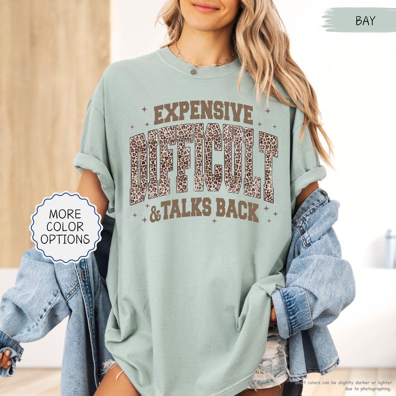 May include: A light green t-shirt with the words "EXPENSIVE DIFFICULT & TALKS BACK" in brown and leopard print lettering. The shirt has rolled-up sleeves and a relaxed fit. The words "MORE COLOR OPTIONS" are in a white circle.