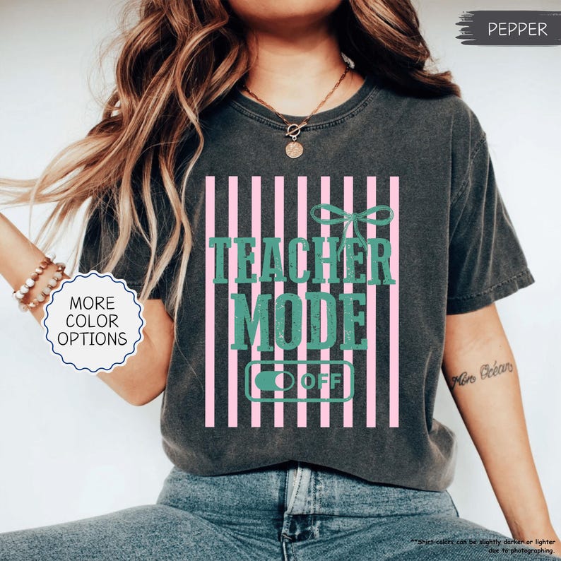May include: Dark gray t-shirt with the text "TEACHER MODE OFF" in green, set against pink vertical stripes. The shirt has a relaxed fit and a crew neck. The words "MORE COLOR OPTIONS" are in a white circle.